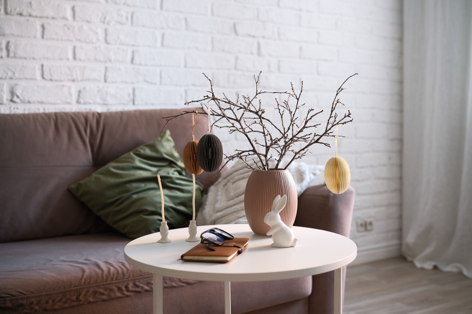 A beautifully cozy living room with modern easter home decor that includes vase with dry branches and eggs, notebook, glasses, ceramic rabbit, candles on coffee table.
